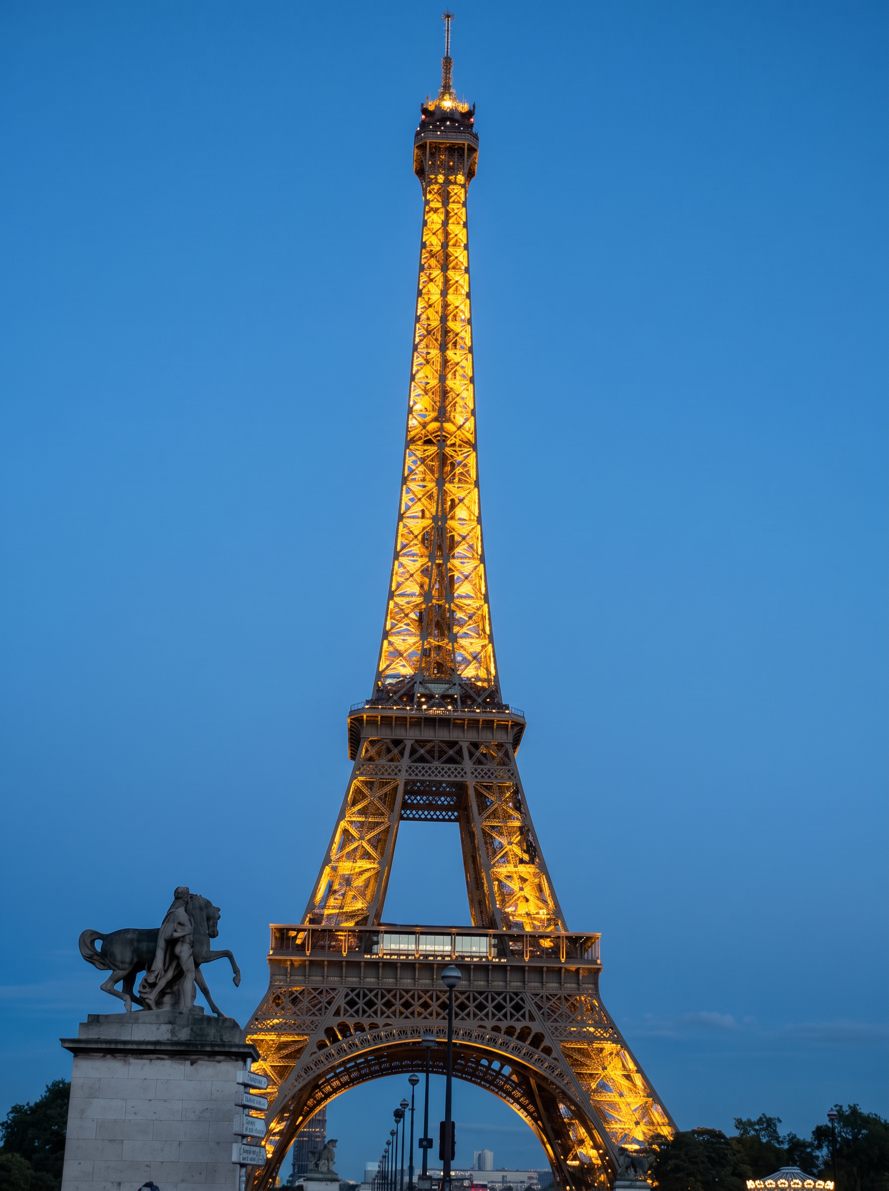 The Eiffel Tower glowing gold against a deep blue twilight sky, with a statue in the foreground